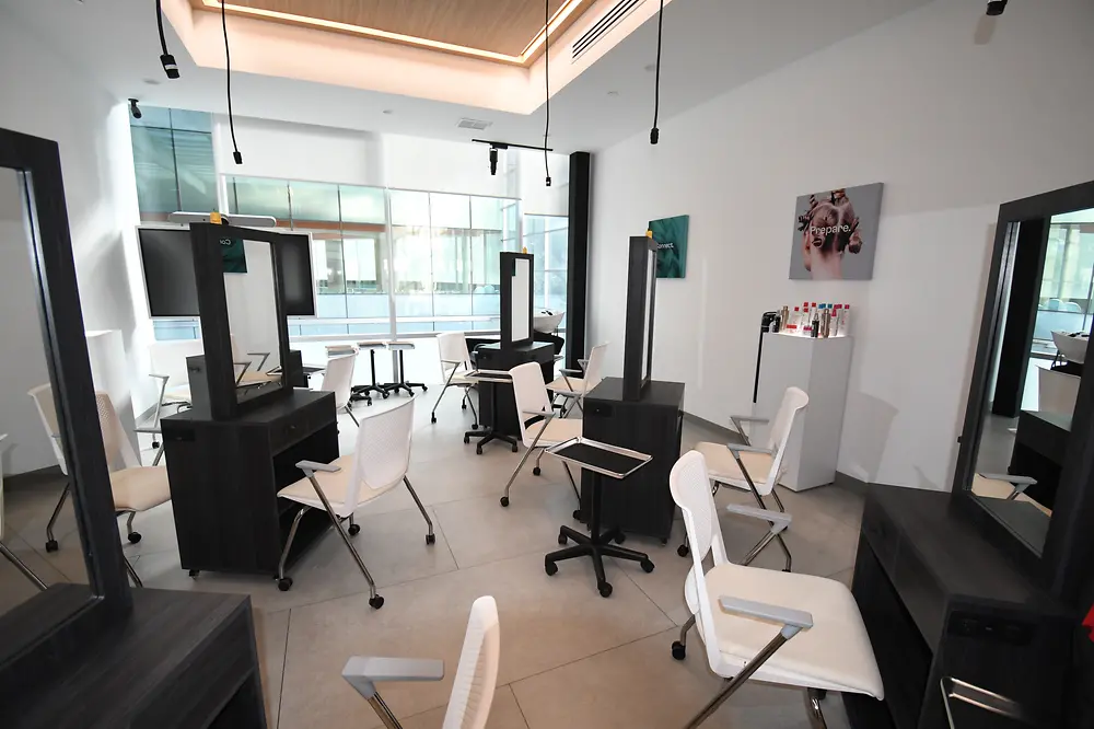 In a bright room, several hairdressing stations are set up, each consisting of a mirror and a white chair.