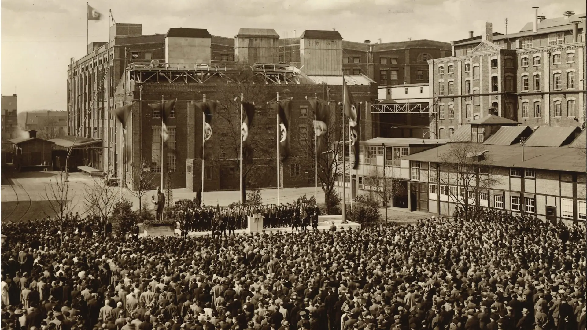 A large crowd gathered in an open square in front of an industrial building. The building is decorated with multiple Nazi flags featuring swastikas, and several flagpoles stand in the center of the square. A group of uniformed individuals is positioned near the flags, while the crowd fills most of the foreground. The surrounding architecture includes multi-story brick buildings and industrial structures.
