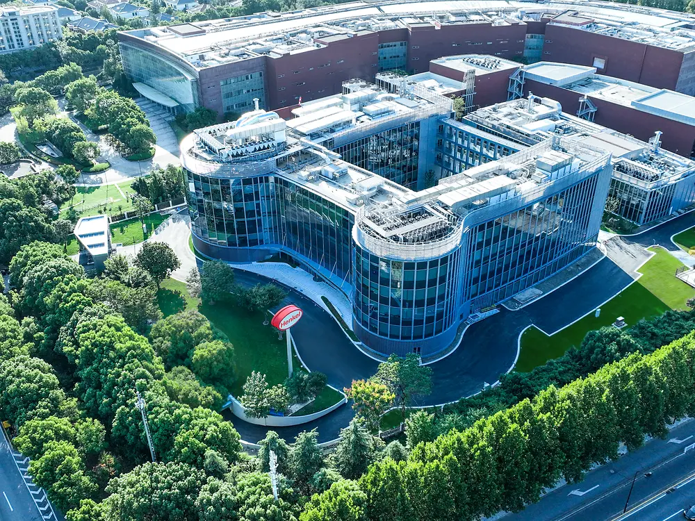 Aerial view of the Inspiration Center Shanghai building surrounded by greenery, with a large red-brick complex behind it.