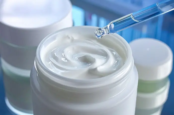 Open cosmetic jar filled with white cream, with other closed jars nearby; a pipette dispensing liquid onto the cream.