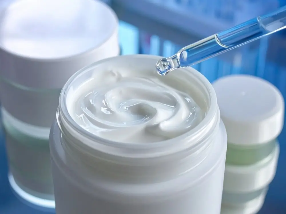 Open cosmetic jar filled with white cream, with other closed jars nearby; a pipette dispensing liquid onto the cream.