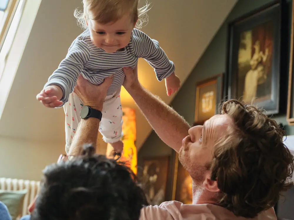 Two fathers holding their baby and smiling at home, symbolizing inclusivity and Henkel’s family-friendly parental leave policy.