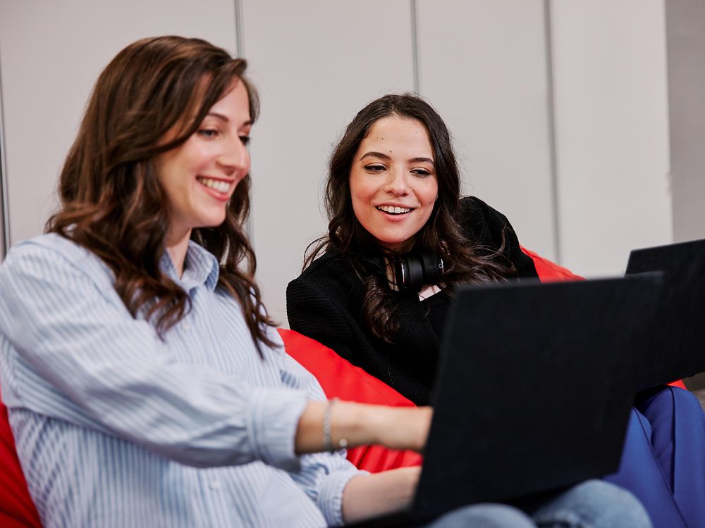 Two people seated on colorful beanbag chairs in a modern office setting, each using a laptop. They appear to be discussing Henkel employee benefits while working collaboratively.