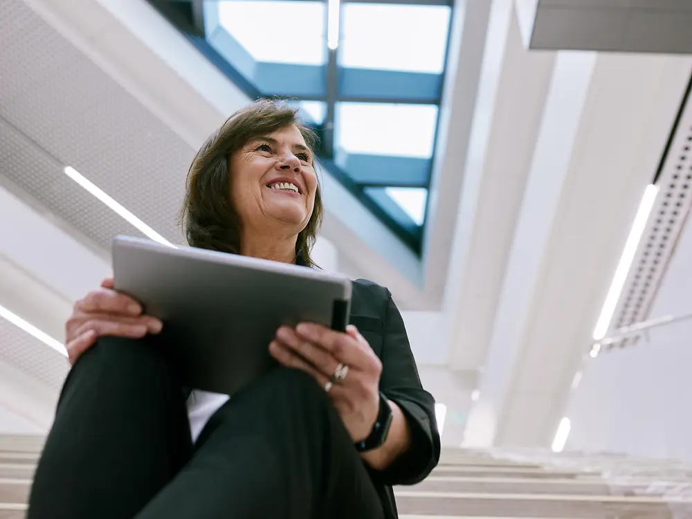 Person sitting on indoor stairs in a modern building, holding a tablet. The background shows a bright, open architectural design with large windows and skylights, creating a clean and professional atmosphere.