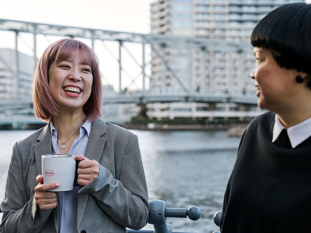Two people having a casual conversation by a waterfront railing, one holding a Henkel-branded coffee mug, with a bridge and city buildings in the background—illustrating Henkel’s flexible work culture.
