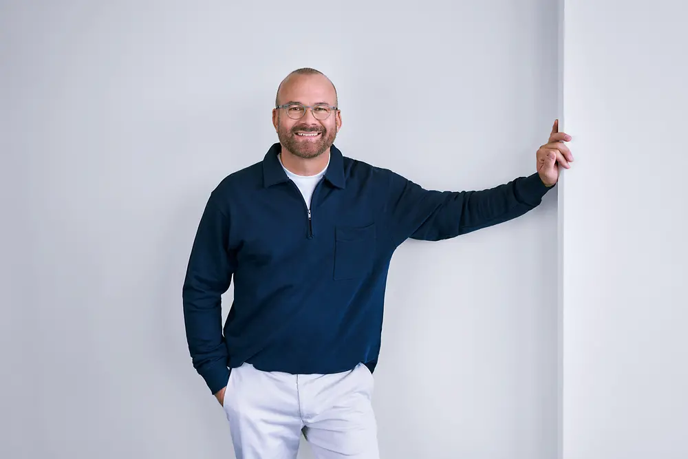 Experienced professional at Henkel, standing confidently against a white wall in a modern setting, wearing a dark long-sleeve shirt and light-colored pants.