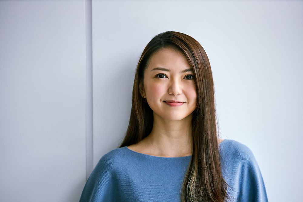Mid-career employee standing against a light-coloured wall, wearing a blue top. The image conveys professionalism and experience, suitable for illustrating career growth and opportunities at Henkel.