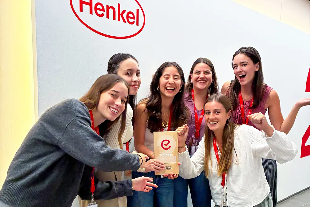 A group of six people is standing together in front of a wall with the Henkel logo. They are wearing red lanyards and casual outfits, and they are gathered closely around an award plaque. The setting appears to be an indoor corporate space with a light-colored wall and gray flooring. The group is posing enthusiastically, highlighting the award in the center.
