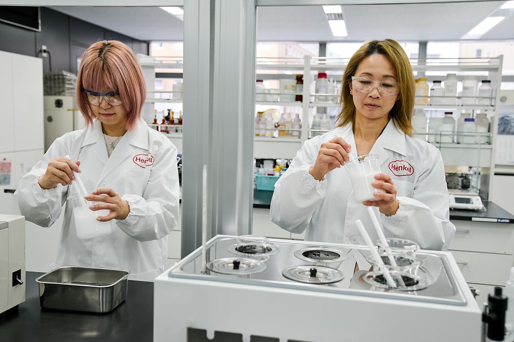Two individuals wearing white Henkel lab coats are working in a modern laboratory. They are handling scientific instruments and containers on a countertop, with various lab equipment and shelves of supplies visible in the background.
