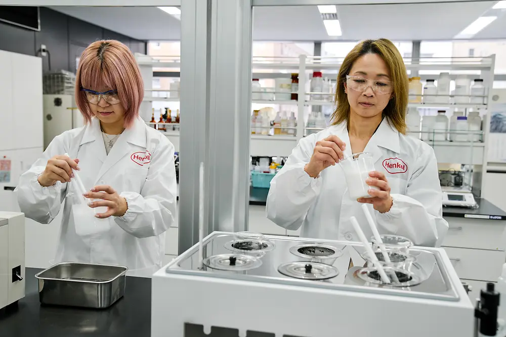 Two individuals wearing white Henkel lab coats are working in a modern laboratory. They are handling scientific instruments and containers on a countertop, with various lab equipment and shelves of supplies visible in the background.