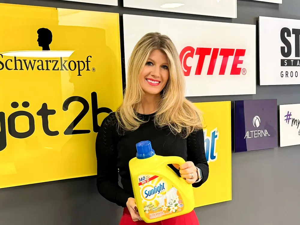 Henkel Employee holding a laundry detergent bottle in front of a wall featuring brand logos such as Schwarzkopf, Purex, and Loctite. Image used for a social media post highlighting how someone crafted a successful career journey within Henkel.