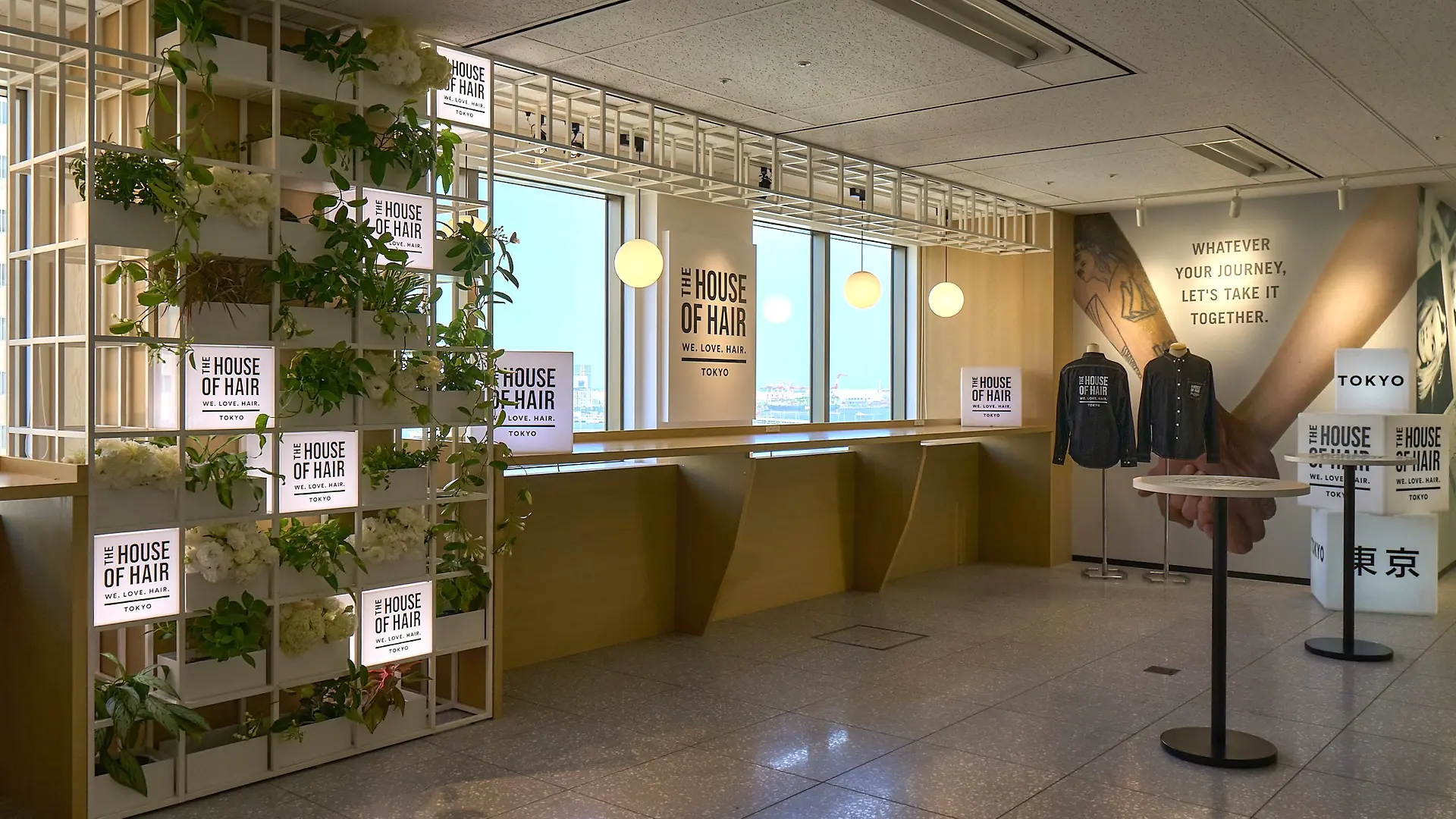 A modern indoor space with large windows and natural light, featuring a wooden counter along the windows and decorative shelving with green plants and illuminated signs reading “House of Hair Tokyo”. On the right side, there is a wall with a motivational quote: “Whatever journey, let’s take it together” and two black hoodies displayed on stands. The overall design is clean and contemporary, with neutral tones and greenery accents.