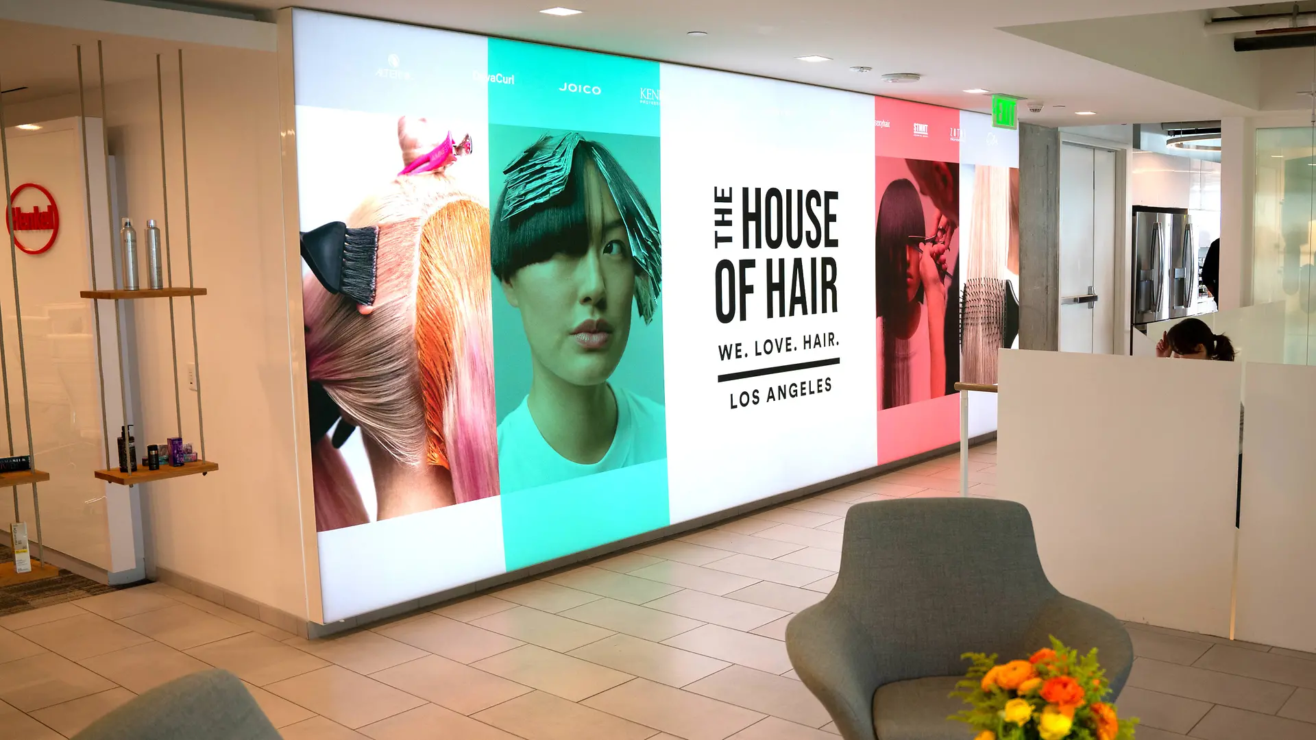 A modern lobby with a bright wall display featuring large panels of colorful hair-themed graphics and the text “THE HOUSE OF HAIR – WE. LOVE. HAIR. – LOS ANGELES.” The space includes light gray lounge chairs, a small table with a vibrant flower arrangement, and a clean, minimalist design with tiled flooring and white partitions.