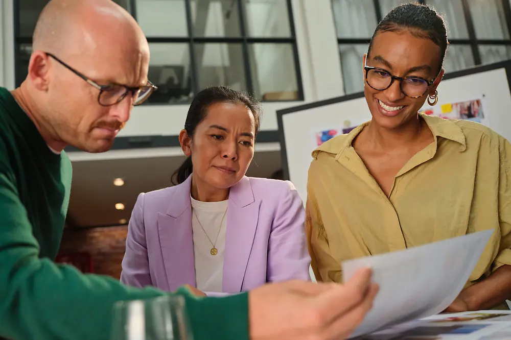 wei Frauen und ein Mann stehen an einem Tisch in einem modernen Büro und betrachten gemeinsam Dokumente.