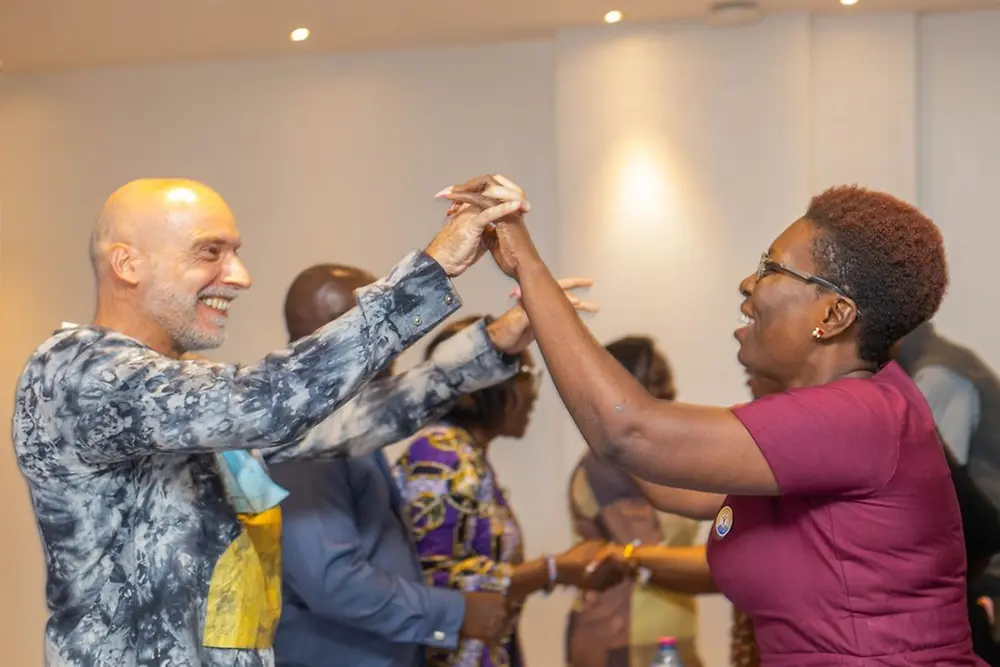 Two people standing indoors, facing each other with their hands interlinked in an upward gesture.