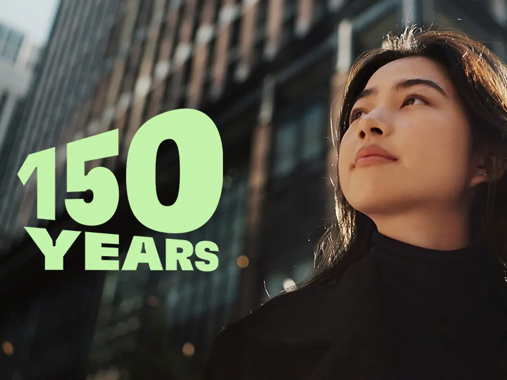 
Green text reading ‘150 YEARS’ over a modern cityscape with tall glass buildings and autumn trees, symbolizing the 150th anniversary celebration.