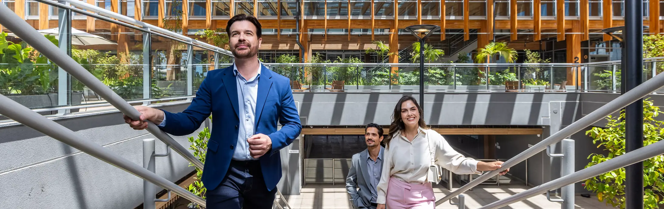 Three people in business attire ascend sunlit stairs within the Inspiration Center Jundiaí, a modern building featuring a glass and wooden ceiling and lush green plants, exuding a positive and collaborative atmosphere.