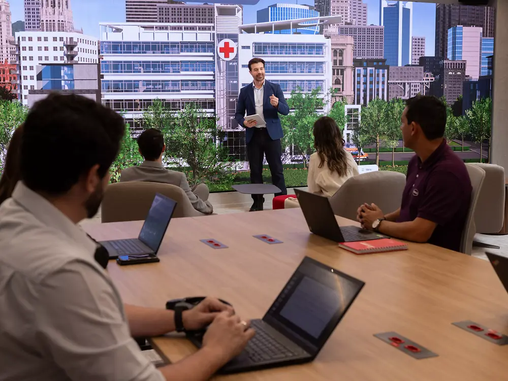 A man presents in front of a digital screen at the Inspiration Center Jundiaí showing a hospital with a red cross. Five people, seated at a table with laptops, listen attentively.