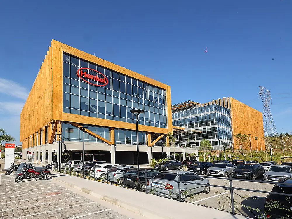 The Inspiration Center Jundiaí, a modern office building with wooden accents and large glass windows, featuring a red Henkel logo. The foreground shows a full parking lot under a clear blue sky.