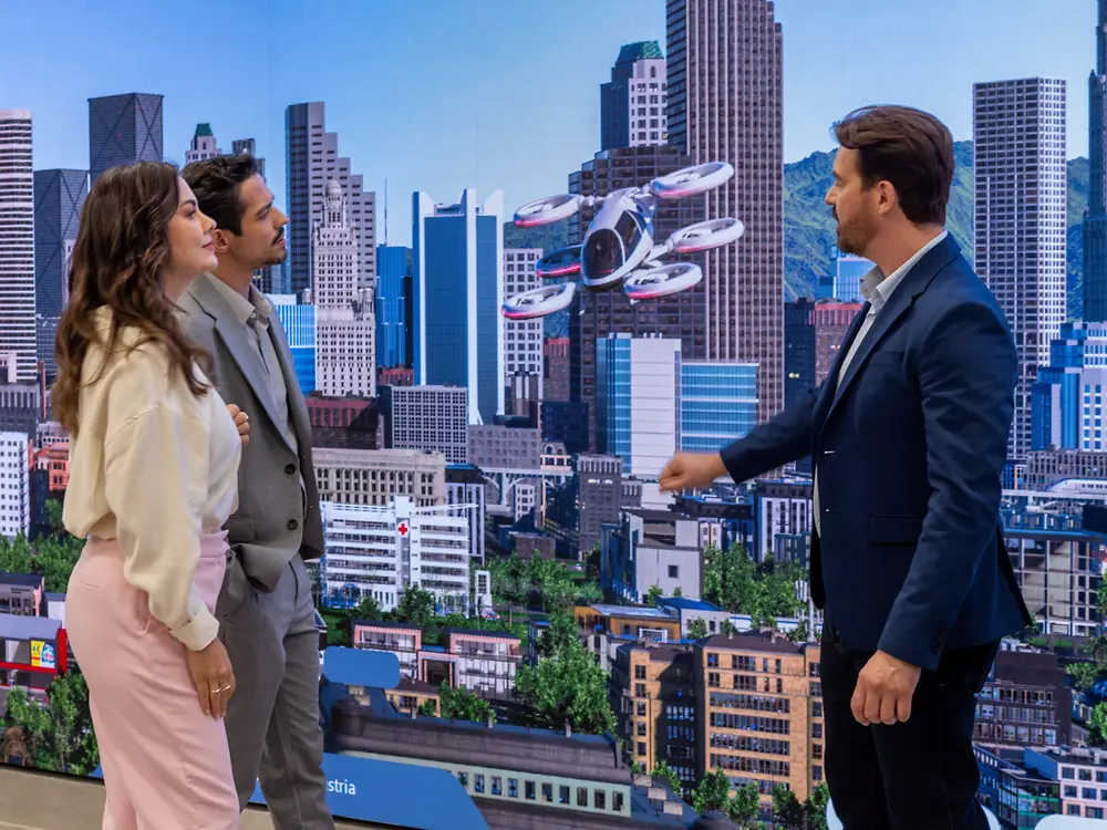 Three people stand indoors at the Infinity Room of the Inspiration Center Jundiaí, facing a large screen showing a futuristic cityscape with flying vehicles. They appear engaged and interested.