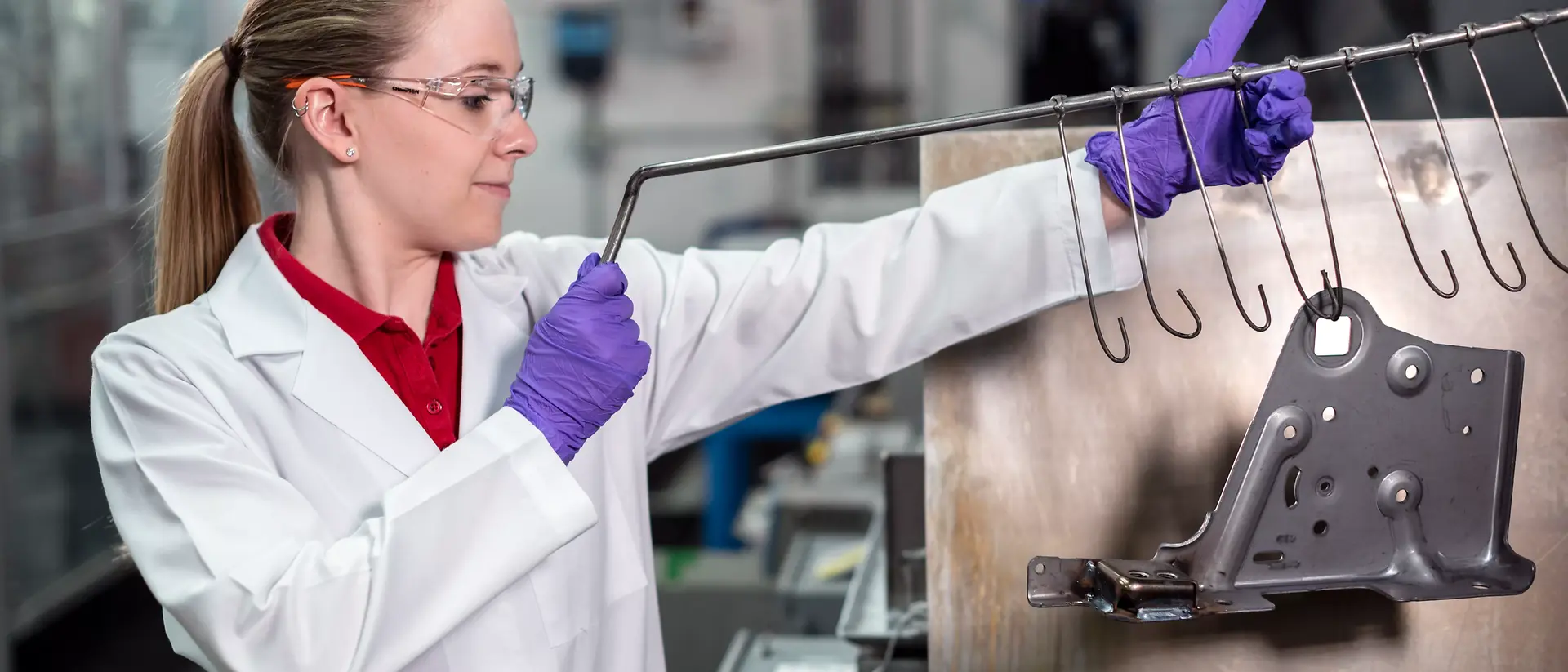 A woman in a lab coat and safety glasses inspects a metal piece on hooks in an industrial setting. She wears purple gloves, conveying focus and professionalism.