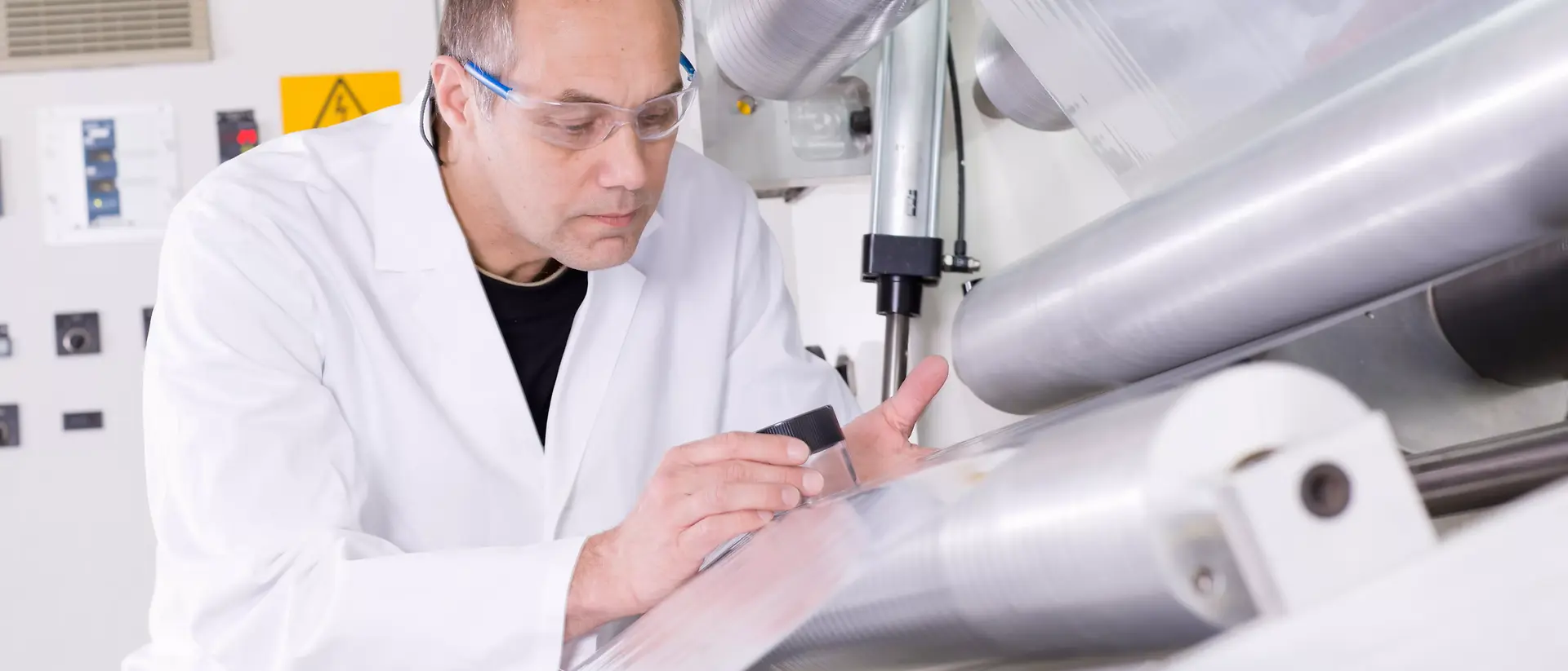 A man in a lab coat inspects a clear plastic sheet on a machine. He wears safety glasses, conveying focus and precision in an industrial setting.