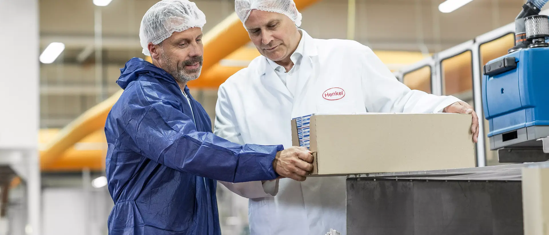 Two men in a production facility, wearing hairnets and protective clothing, inspect a box on a conveyor belt, focusing on quality control for an end of line packaging.