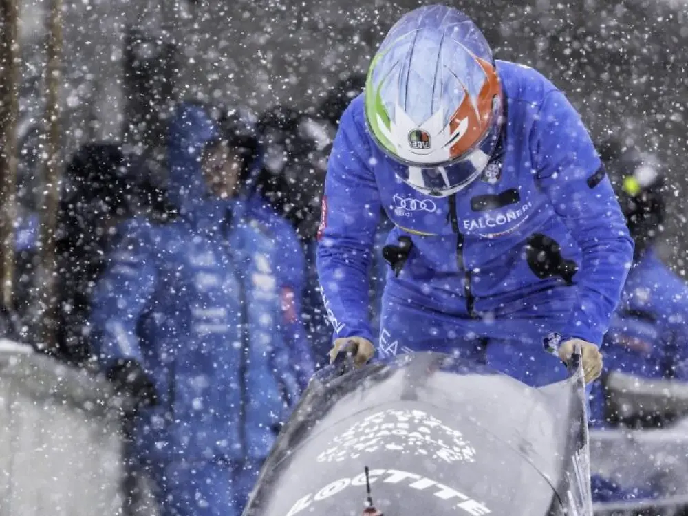 An athlete in a blue uniform pushes a black bobsled forward during heavy snowfall.
