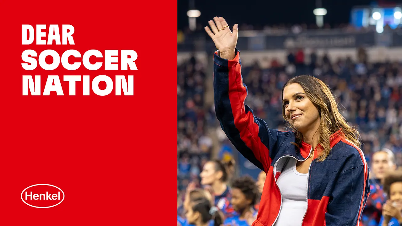 Alex Morgan, a female soccer player waves to the crowd in a stadium while standing with her team, next to a red panel displaying the text “Dear Soccer Nation” and the Henkel logo.
