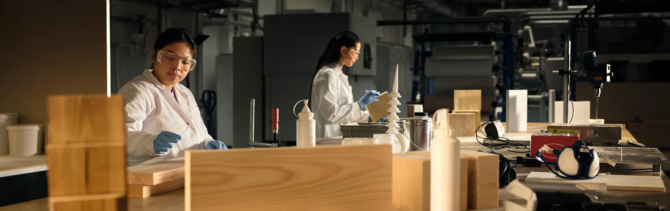 Workshop setting with individuals wearing protective lab coats and gloves, working at a large wooden table covered with wood samples, tools, and bottles.