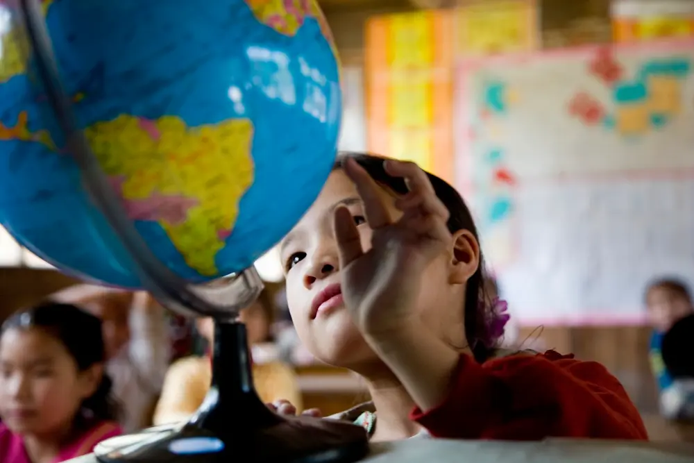 A child in a classroom reaches toward a large globe positioned on a desk, with colorful maps and posters visible on the walls in the background.