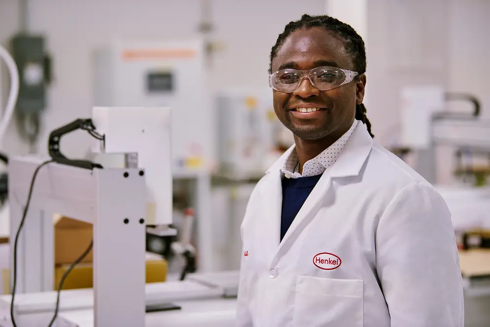 A person in a white Henkel lab coat standing in a laboratory environment surrounded by equipment and machinery.