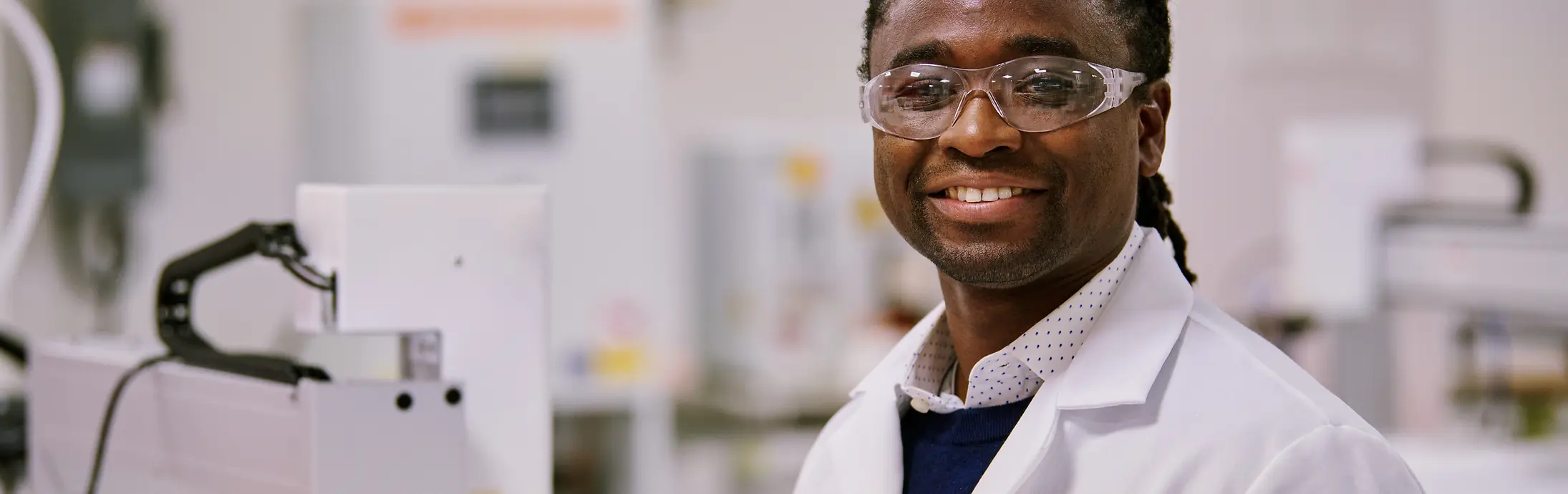 A person in a white Henkel lab coat standing in a laboratory environment surrounded by equipment and machinery.