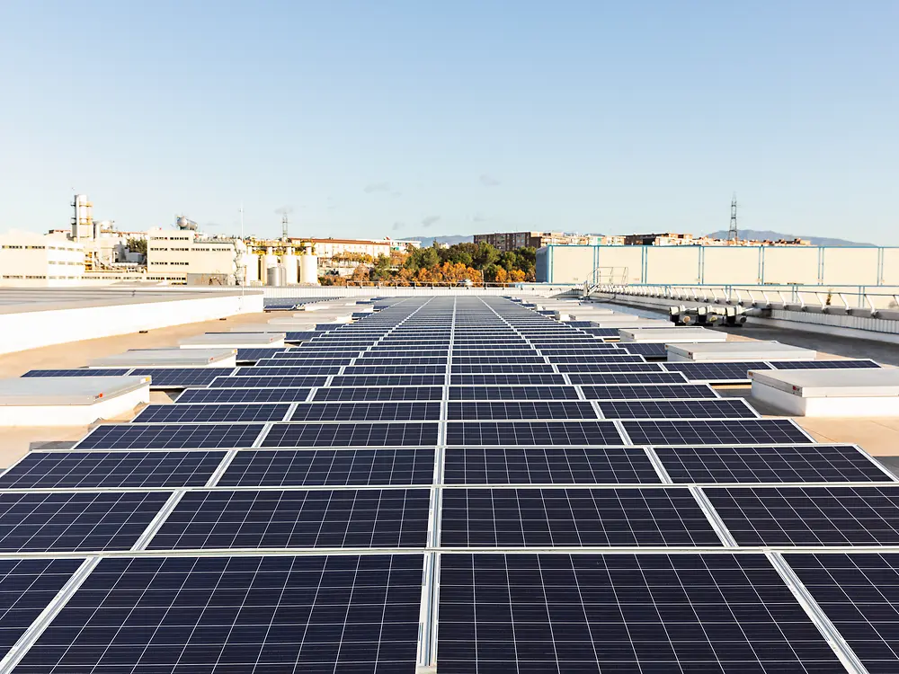 Long rows of solar panels mounted on the flat roof of an industrial building, with factory structures and distant landscape visible in the background on a sunny day.