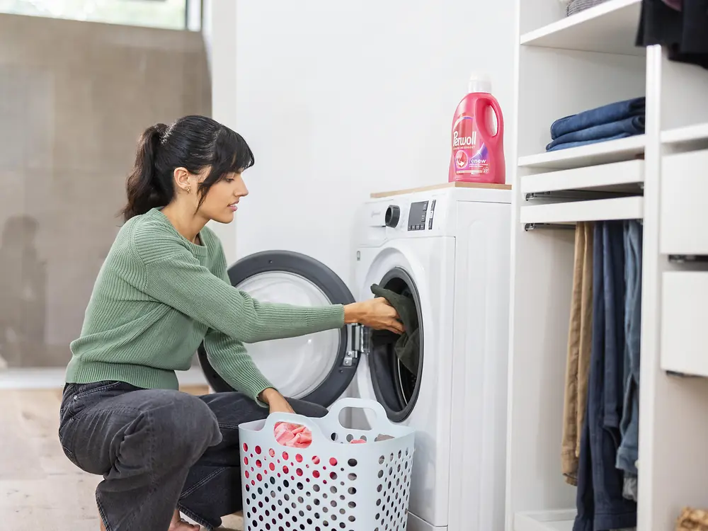 Woman kneeling in front of a front-loading washing machine while placing clothes inside, with a laundry basket on the floor and neatly folded clothes on shelves nearby.
