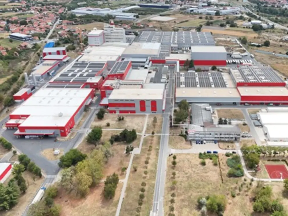 
Aerial view of a large industrial complex with multiple red and white buildings, many of which have roofs covered in solar panels, surrounded by open fields, trees, and nearby residential areas