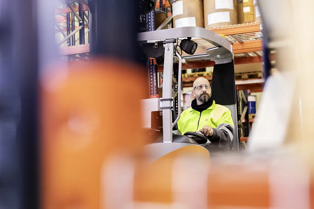 
A warehouse worker wearing a high‑visibility jacket operates a forklift, surrounded by shelves stocked with large containers and equipment