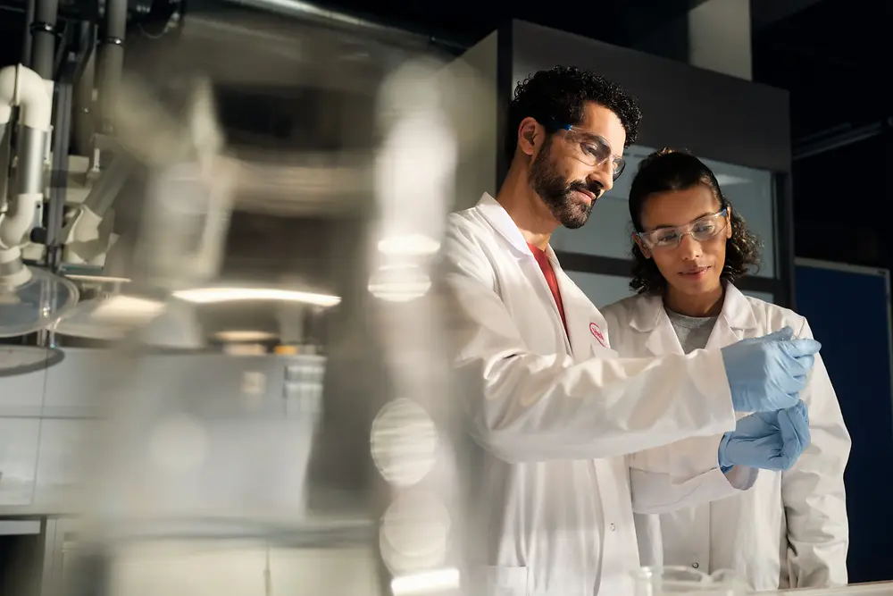 Two scientists wearing white lab coats, safety goggles, and gloves examine a small sample container together in a laboratory setting.