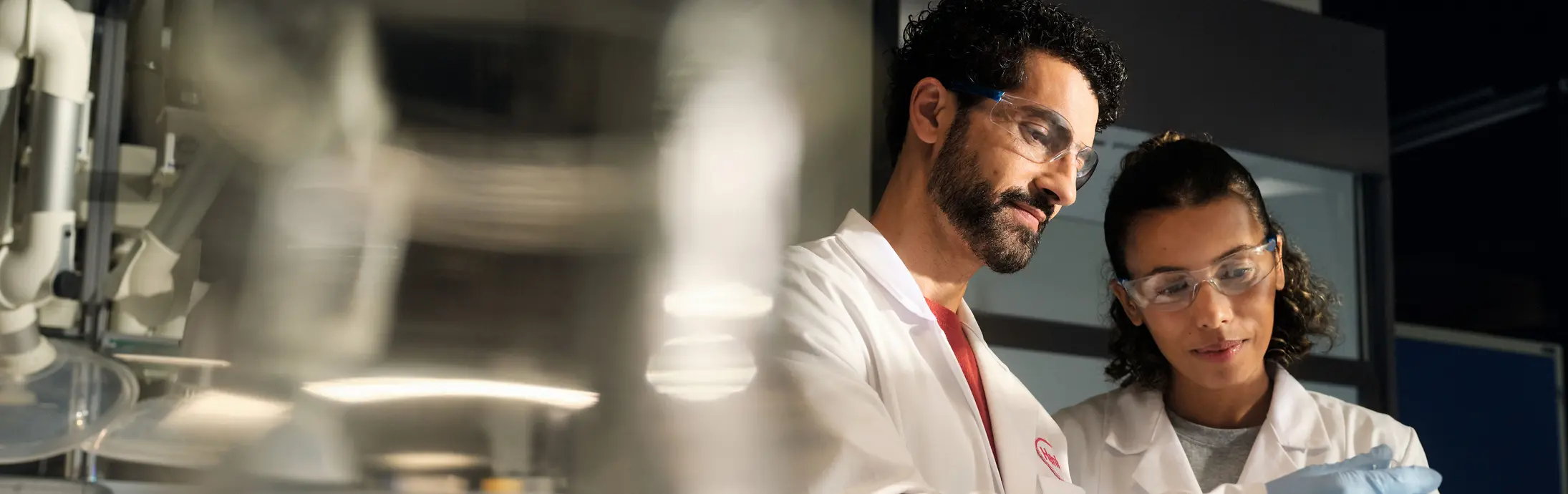 Two scientists wearing white lab coats, safety goggles, and gloves examine a small sample container together in a laboratory setting.