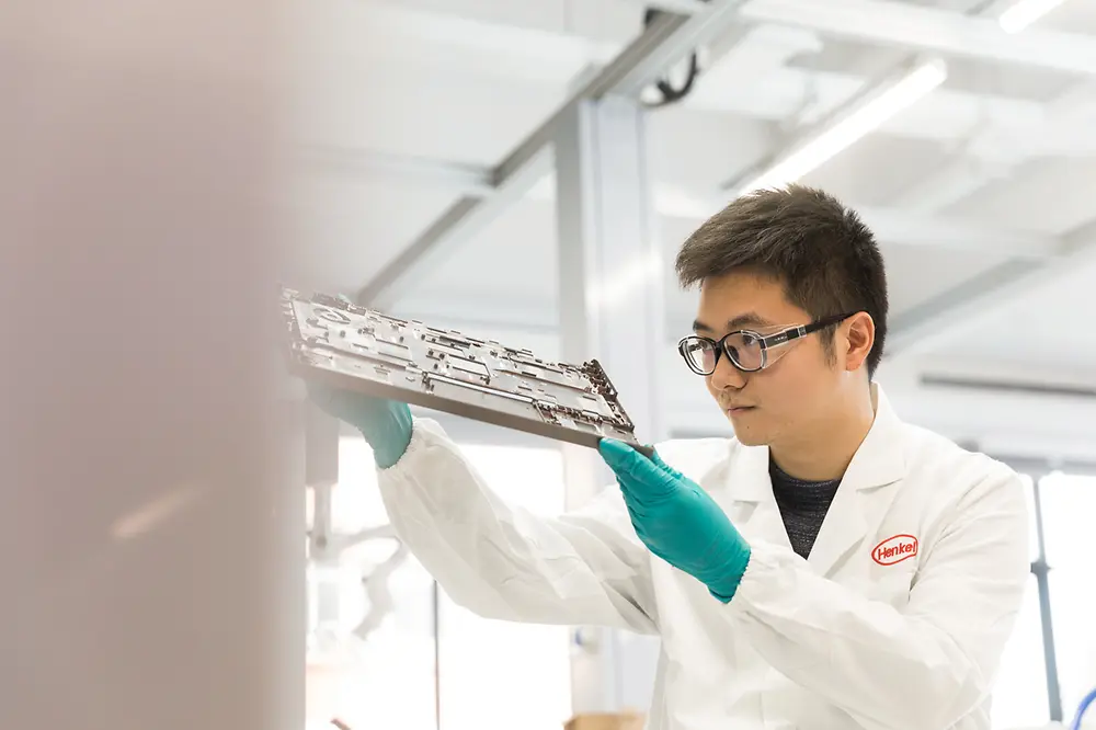 
A technician wearing a white Henkel lab coat and teal gloves closely inspects a metal component in a bright laboratory setting 