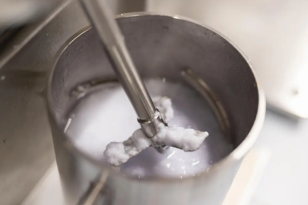 Close-up of a metal container filled with pulped, wet paper fibers being mixed by a mechanical stirrer during a recyclability test.