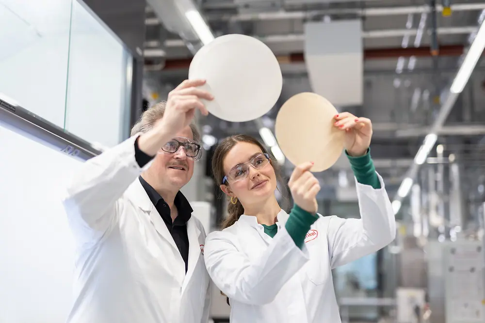 Two researchers in white lab coats and safety glasses stand in a laboratory, examining round paper samples