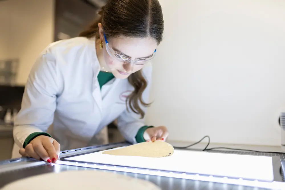 A female researcher in a lab coat examines a round paper sample on a brightly lit inspection table.