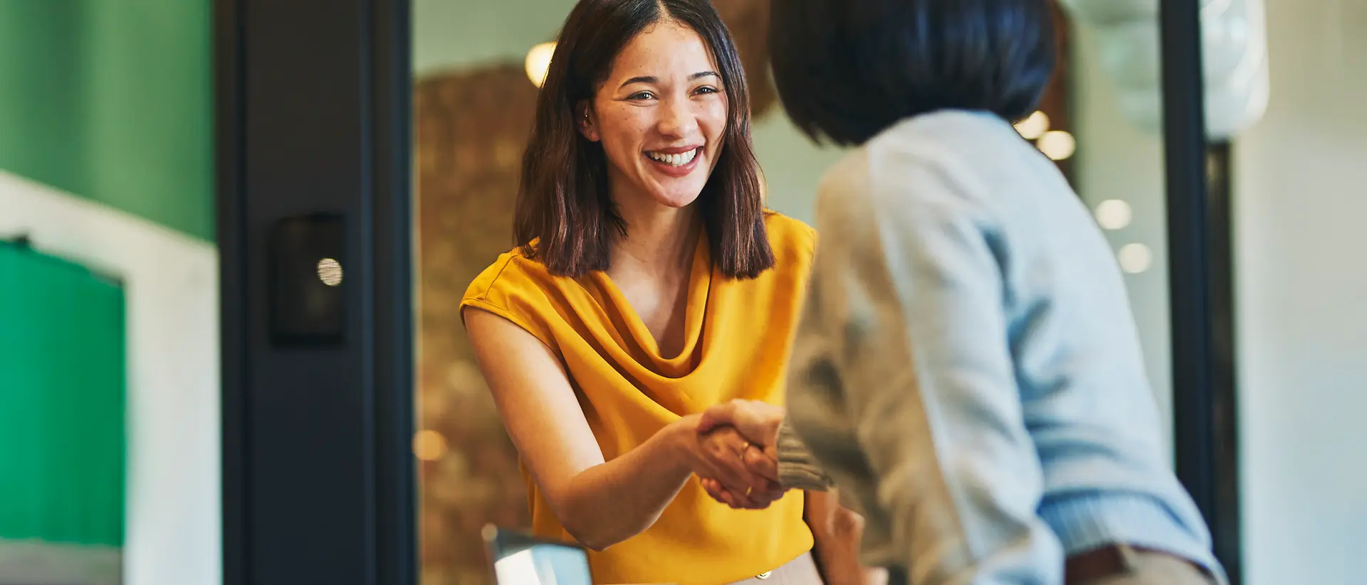 Two people shaking hands across a table in a modern office meeting room, with a laptop visible in the foreground.