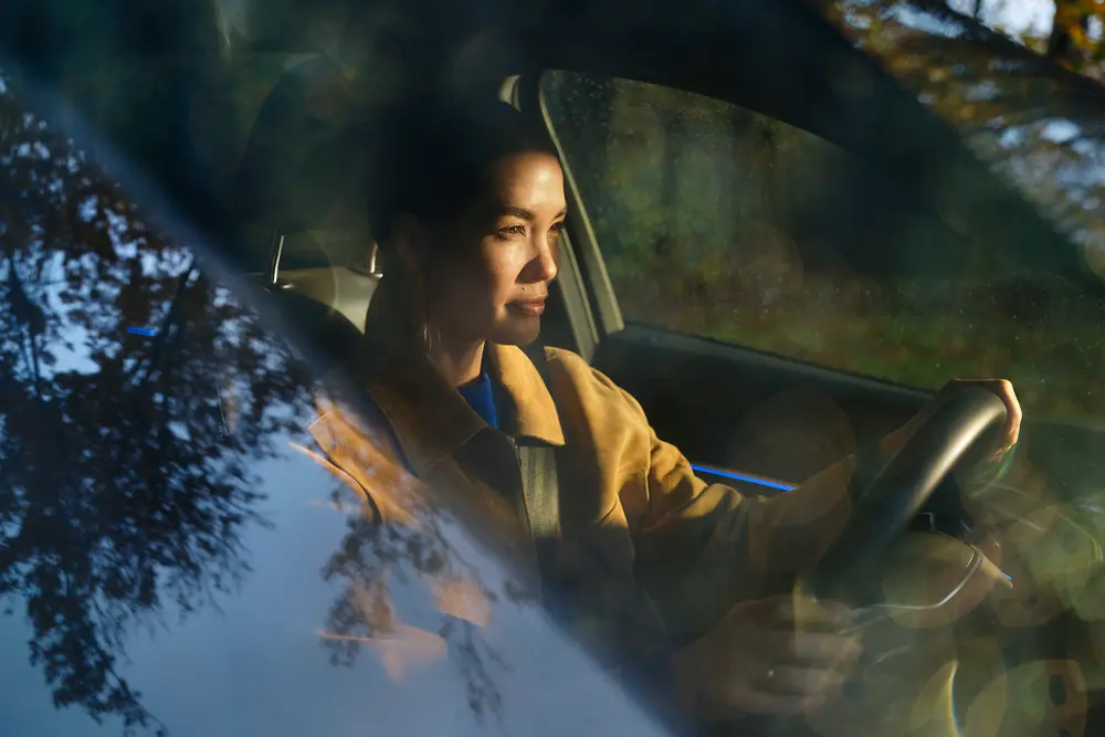 A woman sits in the driver’s seat of a car, photographed through the windshield with reflections of trees on the glass.