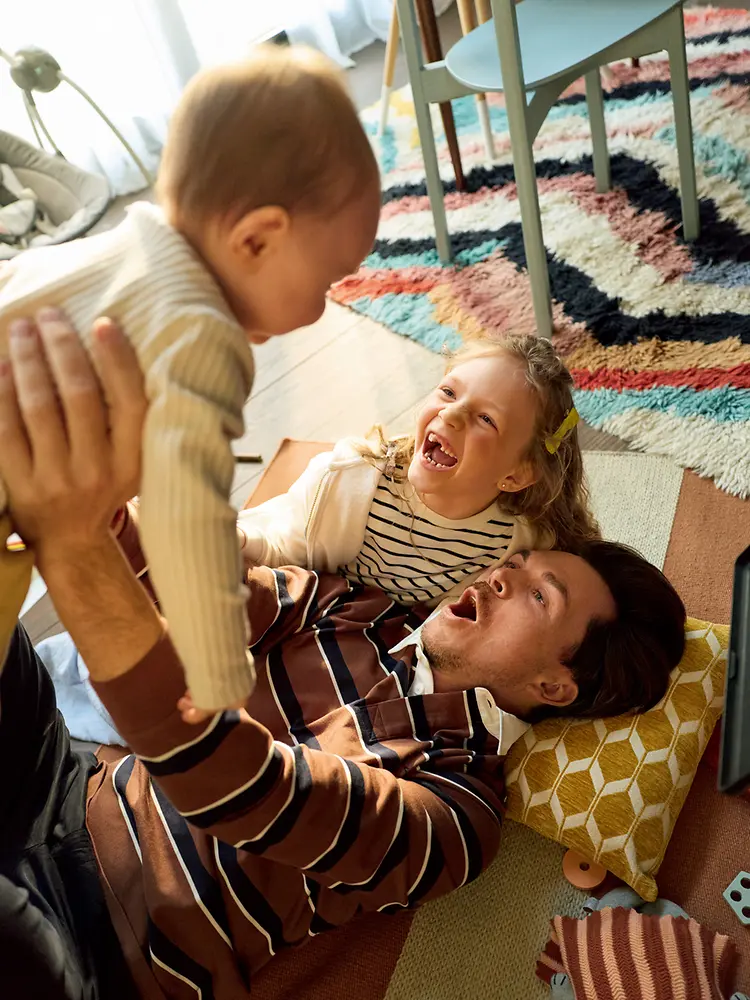 A father is lying on the floor in a living room holding a baby above, while his daughter lies next to him. Both are looking toward the baby.