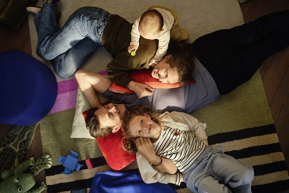 A family consisting of a father, mother, daughter, and baby is lying closely together on rugs and cushions in a living room. The baby sits on one parent while the other family members lie nearby. Some toys are visible in the scene.