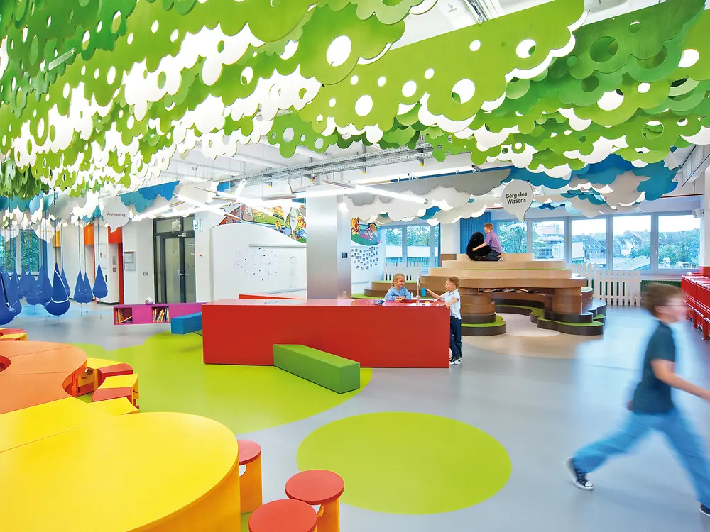 The image shows the Forscherwelt in Düsseldorf, a colorful and modern space with round seating elements and green ceiling decorations.