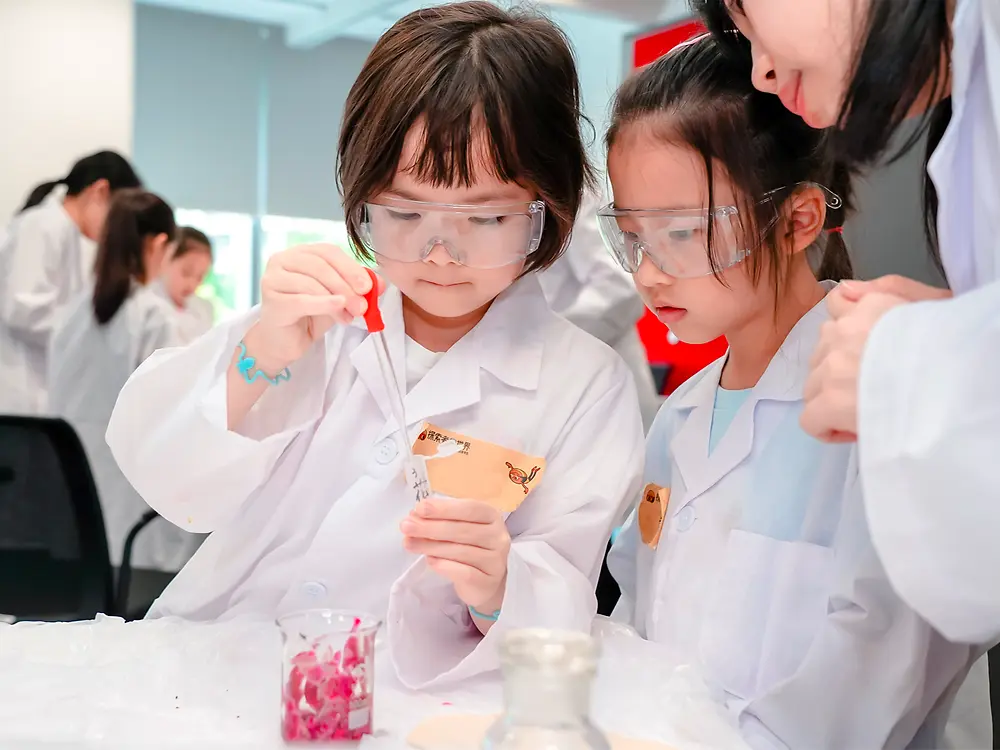 Two children in lab coats and safety goggles stand at a table working with laboratory tools, while an adult observes.