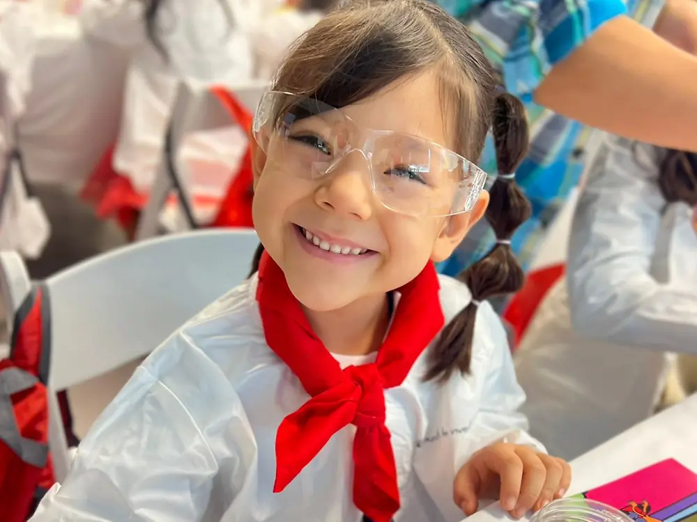 A young child in a lab coat and safety goggles sits at a table with a glass container holding a yellowish liquid.